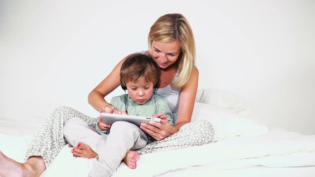 4 j&auml;hriger bub im bett mit kopfh&ouml;rern spielt mit dem tablet gemeinsam mit seiner mutter
