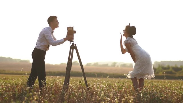 The side view of the happy groom taking photos of the smiling bride in the white dress and black hat using the vintage camera in the field during the sunset.