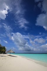 Straw umbrella on the white sandy beach on the coast of bright turquoise ocean and cloudy sky, Maldives