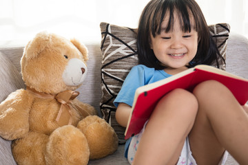 Smiling Asian Chinese little girl reading book with teddy bear