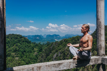 Shirtless male meditating on nature. Young shirtless man sitting on narrow balk in meditation pose on background of amazing tropical views.