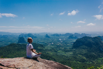 Young male in white meditating on mountain. Man wearing white clothing sitting on high cliff and meditating with breathtaking tropical views on background.