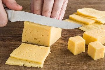 Man's hand with knife cutting a cheese on the wooden board on the table in the kitchen. Dairy product. Healthy eating and lifestyle.