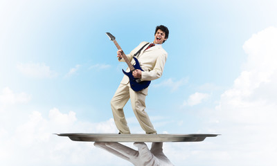 Businessman on metal tray playing electric guitar against blue sky background