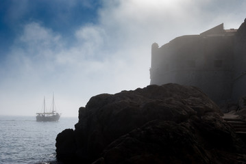 Sailing boat passes the fortified wall of the old city of Dubrovnik on a foggy day