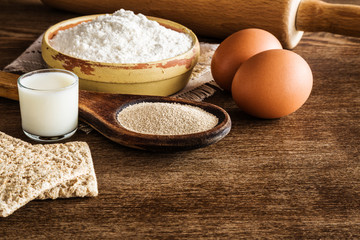 Ingredients for dough on the old wooden table in the kitchen. Yeast in the old wooden spoon. Milk in the glass. Flour in the bowl. Eggs.