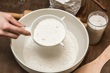 Woman's hand sifting flour through a sieve in the bowl on the old wooden table in the kitchen. Healthy eating and lifestyle.