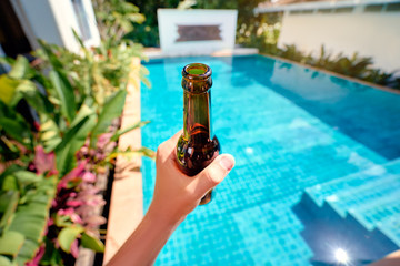 Vacation concept. Male hand holding bottle of beer against swimming pool.