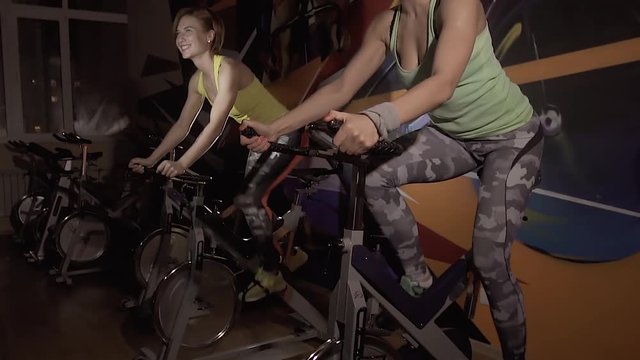 Two active sporty women enjoying their working out on stationary bicycles together