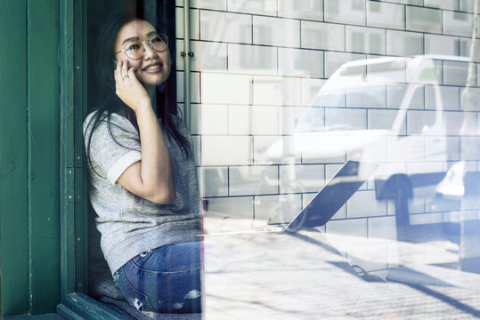 A charming asian travel blogger wearing glasses is talking by phone while sitting in a cafe with the portable computer. Young attractive entrepreneur is making a business call during the coffee break.