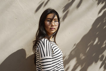 Young asian business woman wearing glasses is looking at the camera while standing beside the bright wall on a street in a sunny day. Student girl is having a walking trip at the city.