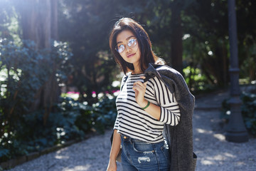 Charming brunette girl is looking at the camera while standing outdoors on the trees and bushes background at the city park. Model look asian woman is walking at the garden on a sunny day.