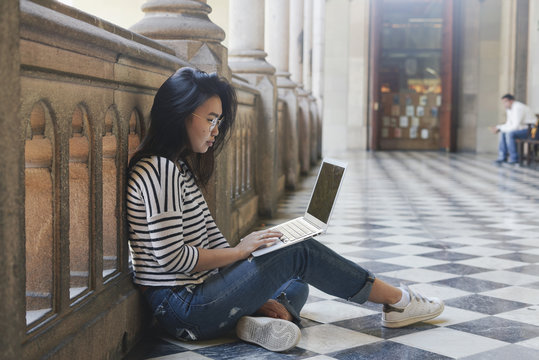 Charming asian student girl wearing glasses is reading email from the friends on the screen of a portable computer connected to the wi-fi while sitting at the balcony of the old city library.