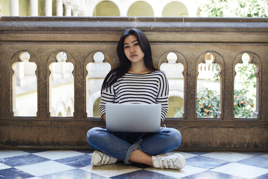 Beautiful model look asian female is looking at the camera while sitting on the floor of the museum balcony with a laptop. Hipster girl is using a portable computer on a university building backgorund