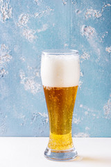 Glass of lager beer with foam on white table with blue texture concrete background.