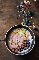 Smoothie bowl healthy breakfast. Chocolate yogurt with blueberries, granola, coconut, lime and carambola over dark wooden texture background. Drawing branch with flowers. Top view, space