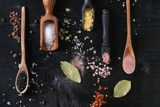 Variety of different colorful salt yellow saffron, pink, black himalayan, white sea and fleur de sel in wooden spoons with black, chili, allspice pepper over black burnt wood background. Top view