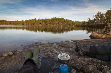 Obraz premium Two persons sitting at the edge of lake making coffe on a camp stove, pine forest in the wilderness of Norway