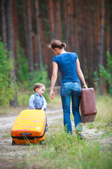 A little boy helps his mom carry suitcases. They together walk along a deserted forest road in a pine forest