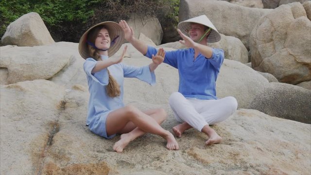 A guy and a girl in Vietnamese hats sit on rocks on the beach in a yoga pose, meditate and look at each other. Concept of a honeymoon date and happiness family, travel in Vietnam