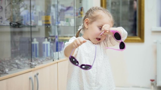 Cute child - blonde Girl dancing and asks mommy to buy colorful glasses in medical store - shopping in clinic