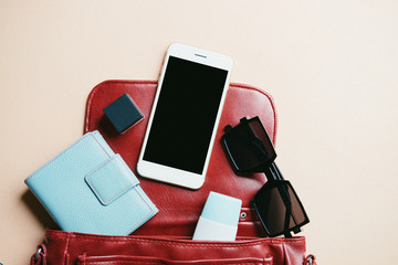 flat lay of red leather woman bag open out with cosmetics, accessories and smartphone on cream color background