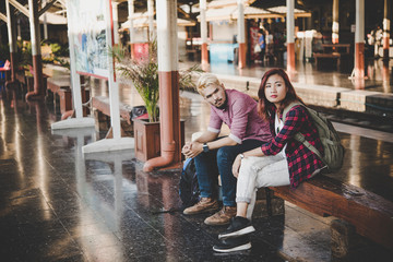 Hipster couple sitting on bench at the train station. Two young tourist are waiting to get on the train and begin their journey. Travel concept.