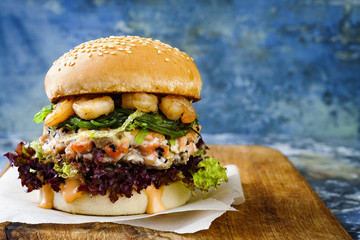 Asian style salmon burger with grilled shrimps, seaweed, lettuce and spicy sriracha mayo sauce served on pieces of brown paper on a rustic wooden board. Bright blue background with copyspace