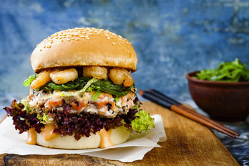 Asian style salmon burger with grilled shrimps, seaweed, lettuce and spicy sriracha mayo sauce served on pieces of brown paper on a rustic wooden board. Bright blue background with copyspace