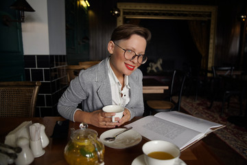 attractive girl in glasses with a short hairstyle in the interior of a stylish cafe reads a book and drinks tea. Intelligent person.