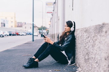 Asian beautiful young woman sitting outdoor listening music with headphones and smartphone hand hold - music, technology, leisure concept