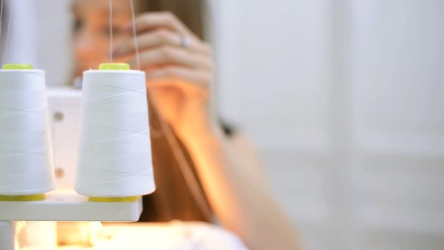 Seamer runs the white thread from the spool into the sewing machine. A woman tailor sits in front of an overlock and prepares it for work on a textile product. At the beginning of the working day, the