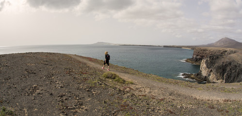 Donna bionda con zaino sulle spalle cammina solitaria sul precipizio di una montagna guardando il mare a Lanzarote, isole canarie