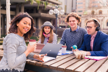 Four positive students sit in time of a break with the folders, with the gadgets, with a globe, with food and one of them listens to music in the earphones in his ears, outdoors