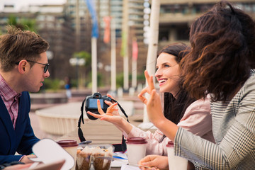 Jolly students sit in time of a break and discuss their tasks with the cups of coffee, with food, with photo camera and one of them shows cool her hand, outdoors