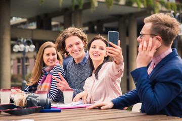 Delighted happy students sit and do selfie-photo on the phone in time of a break, outdoors