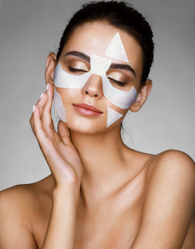 Young sensual lady with cotton facial mask on gray background. Photo of brunette woman touching hand her face. Skin care concept.