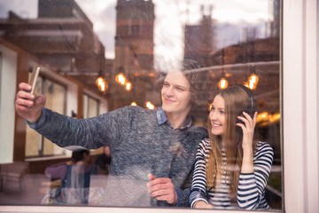 Ecstatic smiling couple sits in cafe and does selfie-photo, a woman listens to music in the earphones on her ears, indoors