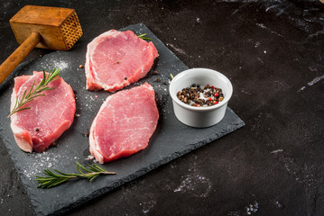 Fresh raw pork, steaks, on a cutting shale board on a black concrete table. Close view, with hammer to beat meat, copy space