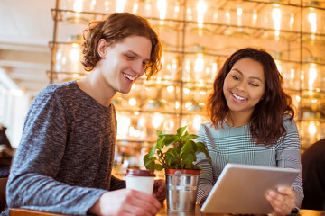 Smiling couple of students sits in cafe and looks at the tablet in time of a break, indoors