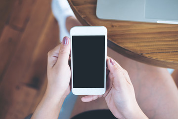 Mockup image of hands holding white mobile phone with blank black screen on thigh with silver color laptop on table and wooden floor background