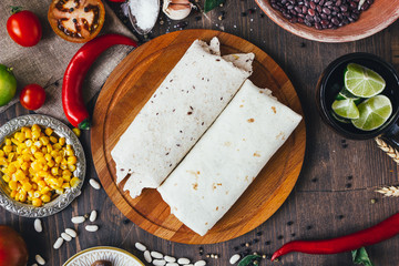 Vegetarian burrito on wooden board over black table surrounded by ingredients.