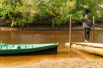 Fisher stands near moored boat on jetty and angles in small river with wooden house on riverside....