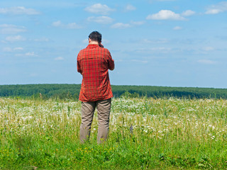 Tall man in bright check shirt stands back before beautiful sunny blossoming meadow with forest in background. Kaluzhsky region, Russia.
