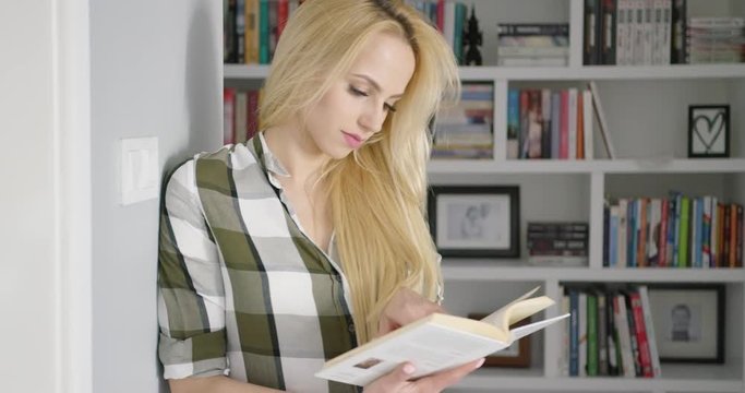 Beautiful young woman wearing checkered shirt and reading book on blurred background of book shelves at home. 
