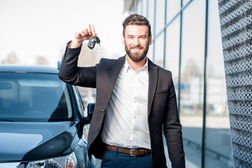 Happy buyer holding keys near the car in front of the modern avtosalon building