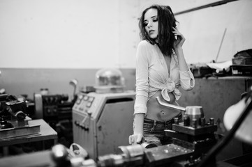 Red haired girl wear on short denim shorts and white blouse posed at industrial machine at the factory.