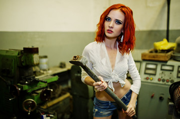 Fototapeta na wymiar Red haired girl wear on short denim shorts and white blouse with hammer at hands posed at industrial machine at the factory.