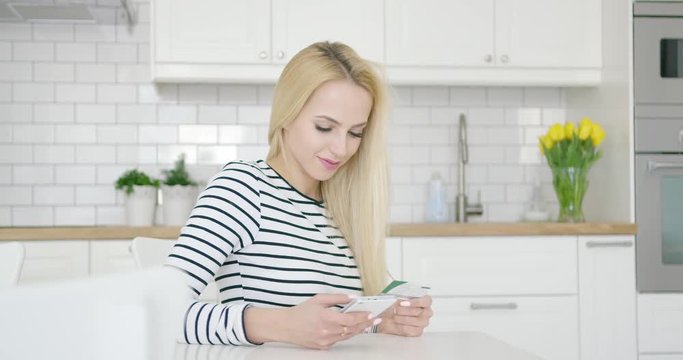 Smiling woman using credit card and phone to buy in Internet while sitting at table in kitchen. 