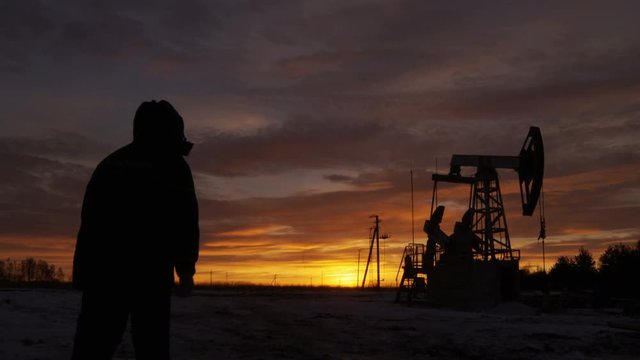 Worker Silhouette in Hood Comes to Pumpjack against Sunset